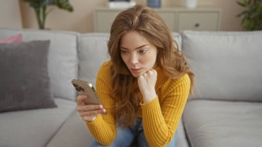 Sarışın, güzel, beyaz bir kadın evdeki oturma odasında oturmuş telefonuna bakıyor, sarı bir kazak giyiyor ve düşünceli bir ifade takınıyor..