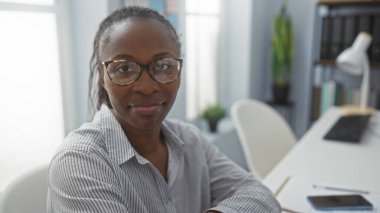 Ofis ortamında çalışan, gözlüklü ve çizgili tişörtlü kendinden emin bir Afrikalı kadın profesyonel tavırlı bir masada oturuyor.