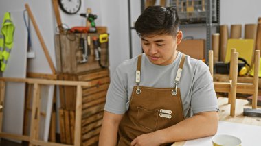 Kahverengi önlüklü genç bir Asyalı, düzenli bir marangozluk atölyesinde oturuyor..