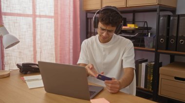 Kulaklıklı genç bir İspanyol, organize bir ofiste dizüstü bilgisayarla çalışırken kredi kartını inceliyor..