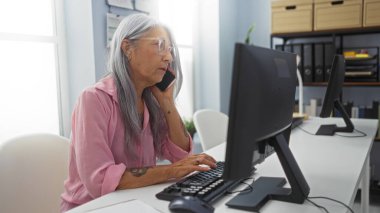 İş yerinde telefonla konuşan bir kadın, ofis ortamında odaklanmış, olgun, gri saçlı birini gösteriyor..