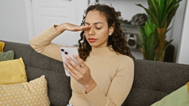 Genç bir kadın, düzgün dekore edilmiş oturma odasındaki kanepede dinlenirken kafası karışmış bir ifadeyle akıllı telefonunu kontrol eder..