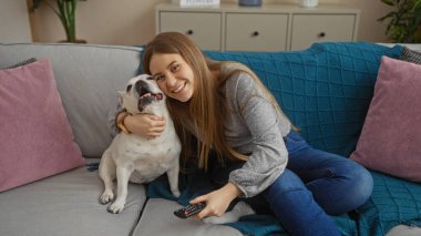 Genç, güzel sarışın bir kadın, sıcak bir oturma odasında oturuyor, elinde uzaktan kumandayla köpeğine sarılıyor..
