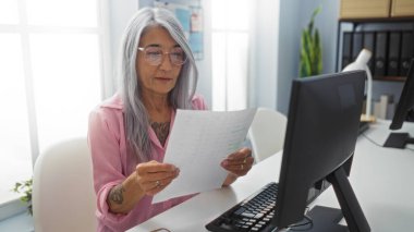 Ofisinde çalışan, masasında gazete okuyan, arka planda bilgisayar, bitki ve raf olan kapalı bir odada gözlük takan bir kadın..