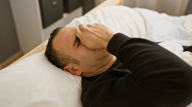 Kapalı bir yatakta yatan stresli bir İspanyol adam. Alnını tutarken sıkıntılı görünüyor. Umutsuzluğu ya da baş ağrısını tasvir ediyor..
