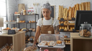 Beyaz saçlı kadın bir pastanenin içinde duruyor. Etrafı ekmek ve hamur işleriyle çevrili. Sıcak ve davetkar bir dükkan ortamında..