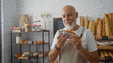 Orta yaşlı, gri saçlı ve sakallı bir adam sıcak bir pastanenin içinde taze fırında ekmek tutuyor..