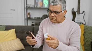 Mature man in living room examining medicine bottle while holding smartphone, with guitar in background. clipart