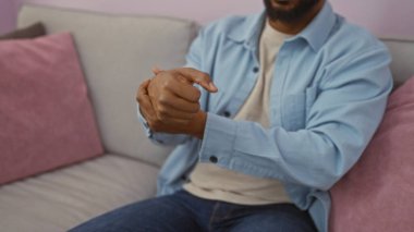 Kanepede pembe ve gri yastıklı rahat bir oturma odasında otururken bileğini acı içinde tutan sakallı genç bir adam.