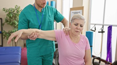 İyi donanımlı bir rehabilitasyon odasında erkek bir terapistle omuz fizyoterapisi gören bir kadın hasta..