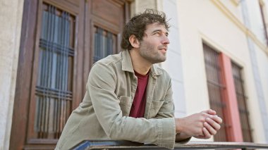 Yakışıklı, sakallı genç bir adam güneşli bir şehir sokağında korkuluklara yaslanıyor..