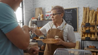 Kadın fırının içinde ekmek ve hamur işleriyle erkeklere hizmet ediyor. Tahta raflardaki baget de dahil. Kapalı bir dükkanda iki yetişkin işçinin takım çalışmasını sergiliyor..