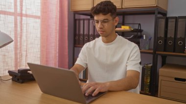 Ofiste dizüstü bilgisayarda çalışan İspanyol bir adam. Arka planda dosyalar ve kutular var. Profesyonel bir çalışma alanını gösteriyor..