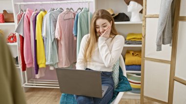 Renkli bir odada dizüstü bilgisayar kullanan gülümseyen genç sarışın bir kadın. Giysileri ve mankeni var. Sıradan moda ve yaşam tarzını somutlaştırıyor..