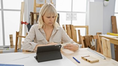 A focused blonde woman using a tablet in a bright carpentry workshop full of wooden furniture and tools. clipart