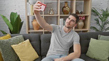 Yakışıklı İspanyol bir adam akıllı telefonuyla selfie çekiyor, modern oturma odasındaki gri bir kanepede oturuyor..