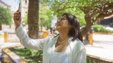 Orta yaşlı, kısa saçlı bir İspanyol kadın güneşli bir şehir parkında gözlük ve beyaz gömlek giyerken geniş bir gülümsemeyle selfie çekiyor..