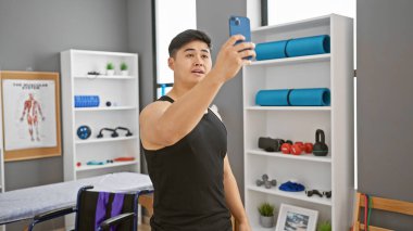 Young asian man taking a selfie in a modern rehabilitation clinic's gym, with exercise equipment in the background. clipart