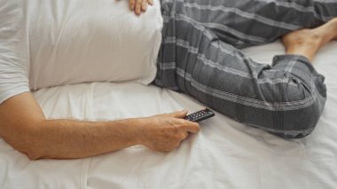 Orta yaşlı İspanyol bir adam yatak odasında uzanıp elinde uzaktan kumandayla evde ekose pijama giyiyor.
