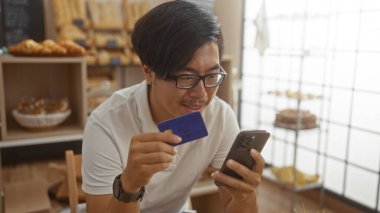 Genç adam elinde kredi kartı tutarken fırının içinde akıllı telefon kullanıyor ve sergilenen taze ekmeği var.