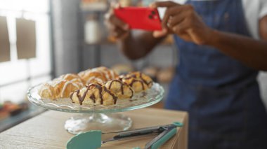 Genç adam fırında kırmızı telefon kullanarak hamur işlerini fotoğraflıyor. Çikolatalı kruvasanları ve cam tezgahın üzerindeki kremayı vurguluyor.