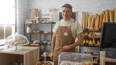 Bir fırında duran genç adam çeşitli ekmek ve hamur işleri sergiliyor, kahverengi önlük giyerken güvenle gülümsüyor.