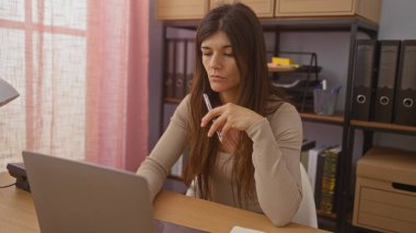 Ev ofisinde çalışan, dizüstü bilgisayarına odaklanmış ve elinde kalemle arka planda raflar ve dosyalarla bir masada oturan çekici İspanyol bir kadın..