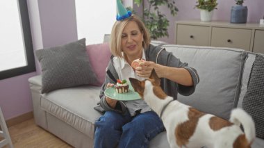 Mature woman celebrating a special occasion at home, interacting with her adorable dog in the living room, while wearing a party hat and holding a plate with a mini cake. clipart