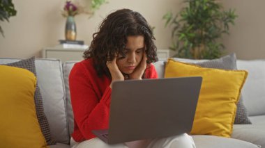 Esmer saçlı genç bir İspanyol kadın oturma odasında oturuyor ve dizüstü bilgisayarla çalışıyor. Stresli ve düşünceli görünüyor. Etrafında rahat yastıklar ve bir apartman dairesinde kapalı bitkiler var..