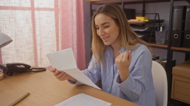 Genç sarışın kadın, arka planda raflar olan ofis ortamındaki bir masada mutlu bir ifadeyle bir belge okuyor..