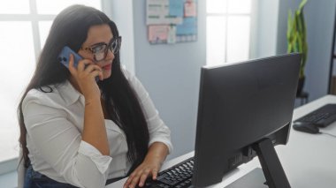 Gözlüklü genç bir kadın telefonda konuşurken bilgisayar başında çalışıyor. Parlak ışıklandırması ve düzenli çalışma alanı olan bir ofiste.