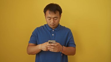 Genç, yakışıklı, Asyalı bir adam mavi tişörtlü, izole edilmiş sarı arka plan duvarına karşı akıllı telefon kullanıyor.
