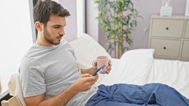 Genç bir İspanyol adam, rahat bir yatak odasının içinde yatarken telefonunu ve fincanını tutuyor..