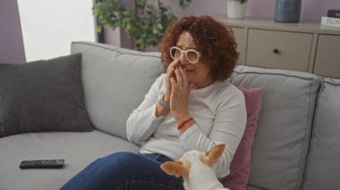 Orta yaşlı bir kadın ve evcil köpeği, rahat bir oturma odasında, ev dekoruyla çevrili, koltukta duygusal görünen bir kadınla oturuyor..