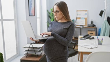 Gözlüklü hamile kadın modern ofis ortamında dizüstü bilgisayar kullanıyor, iş yerinde annelik gösterisi yapıyor..