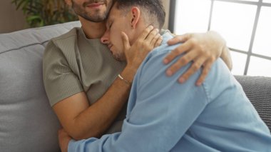 Oturma odasındaki bir kanepede sevgiyle kucaklaşan, sıcak bir yuva ortamında sevgi ve şefkat ifade eden, lgbtq ailesini ve ilişki dinamiklerini ön plana çıkaran iki adam..