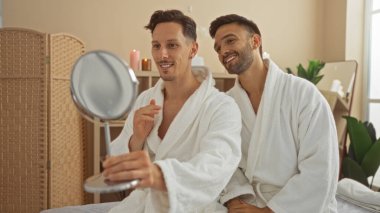 Beyaz cüppeli iki adam sıcacık bir spa odasında aynaya bakıp gülümserken, lgbtq dostu bir sağlık merkezinde aşkın ve rahatlığın özünü yakalıyorlar.