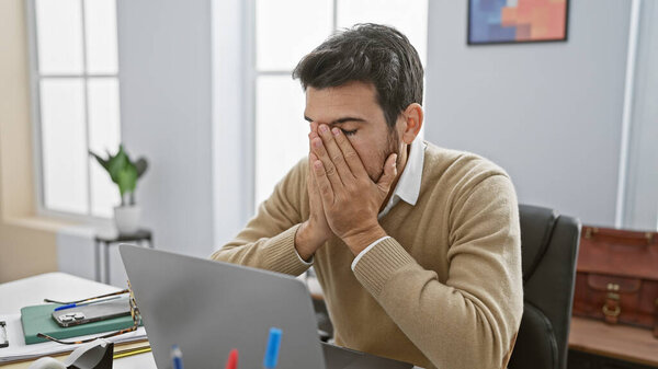 A stressed hispanic man with a beard covers his face in a modern office setting, suggesting exhaustion or headache.
