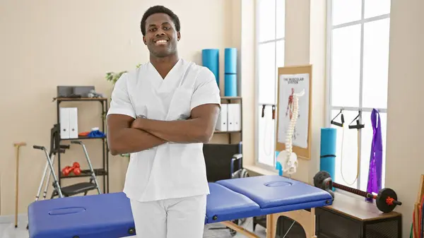 stock image Smiling african american male healthcare professional with arms crossed standing in a modern rehab clinic interior.