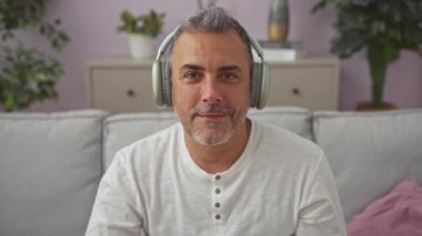 Yakışıklı, orta yaşlı, kulaklıklı bir adam oturma odasındaki koltukta otururken gülümseyen kulaklıklarla müzik dinleyip, rahat ve neşeli bir ev atmosferi yaratıyor..