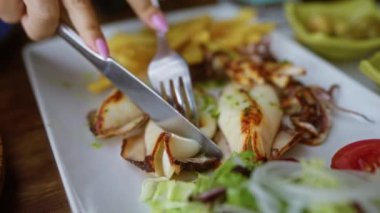 Restoranda beyaz bir tabakta çatal ve bıçakla ızgara kalamar dilimleyen bir kadın.