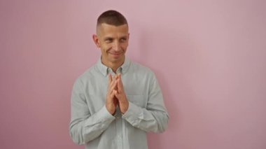 İzole edilmiş pembe arka planda duran genç adam, elleri sımsıkı bağlanmış, açık tişörtlü ve hafif bir gülümseme sergiliyor.