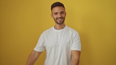 Beyaz tişörtlü genç İspanyol adam yan yan duruyor ve gülümsüyor, iki eliyle ve parmaklarıyla kamerayı izole edilmiş sarı arka planda gösteriyor.