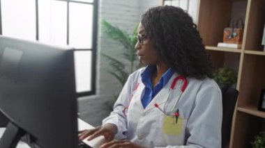 Bilgisayarında çalışan ve klinik ortamında telefonla konuşan kıvırcık saçlı, gözlüklü ve steteskoplu Afro-Amerikan kadın doktor..