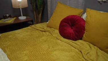 Yatak odasındaki Afrikalı Amerikalı genç bir kadın kırmızı yastıklı hardal sarısı bir yatakta, yanında lamba ve dekor eşyaları olan bir komodinin yanında yatıyor..