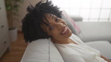 Güzel, genç, Afro-Amerikan bir kadın kıvırcık saçlı sıcak bir şekilde gülümserken rahat, aydınlık bir oturma odasında kanepede dinleniyor..