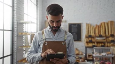 Arka planda ekmek ve hamur işleri sergilenen bir pastanede tablet kullanan sakallı genç adam.
