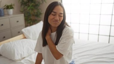 Boyun ağrısı çeken bir kadın şık bir yatak odasında beyaz bir yatakta oturur ve arka planda çiçekleri, rahat bir ev içinde günlük beyaz bir gömlek giyer..