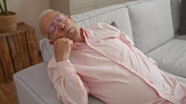 Gözlüklü yaşlı bir adam oturma odasındaki kanepede pembe bir gömlek giyerek uyur. Huzurlu ve huzurlu bir iç mekan ortamı yaratır..
