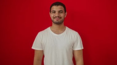 Beyaz tişört giyen genç İspanyol adam gülümsüyor ve olumlu bakıyor. Kırmızı arka plandan onay almak için başparmağını kaldırıyor.